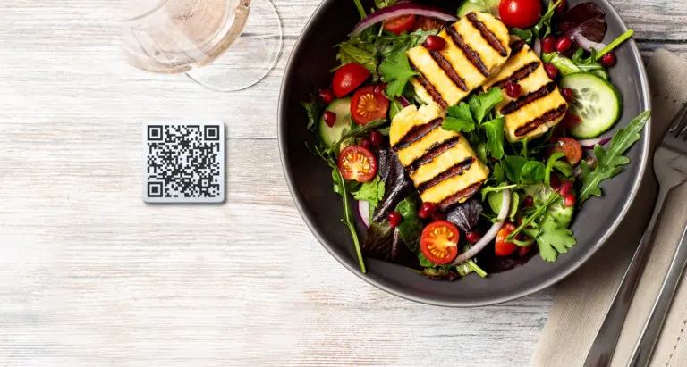Bowl of grilled halloumi salad on a wooden table next to a QR code table tag