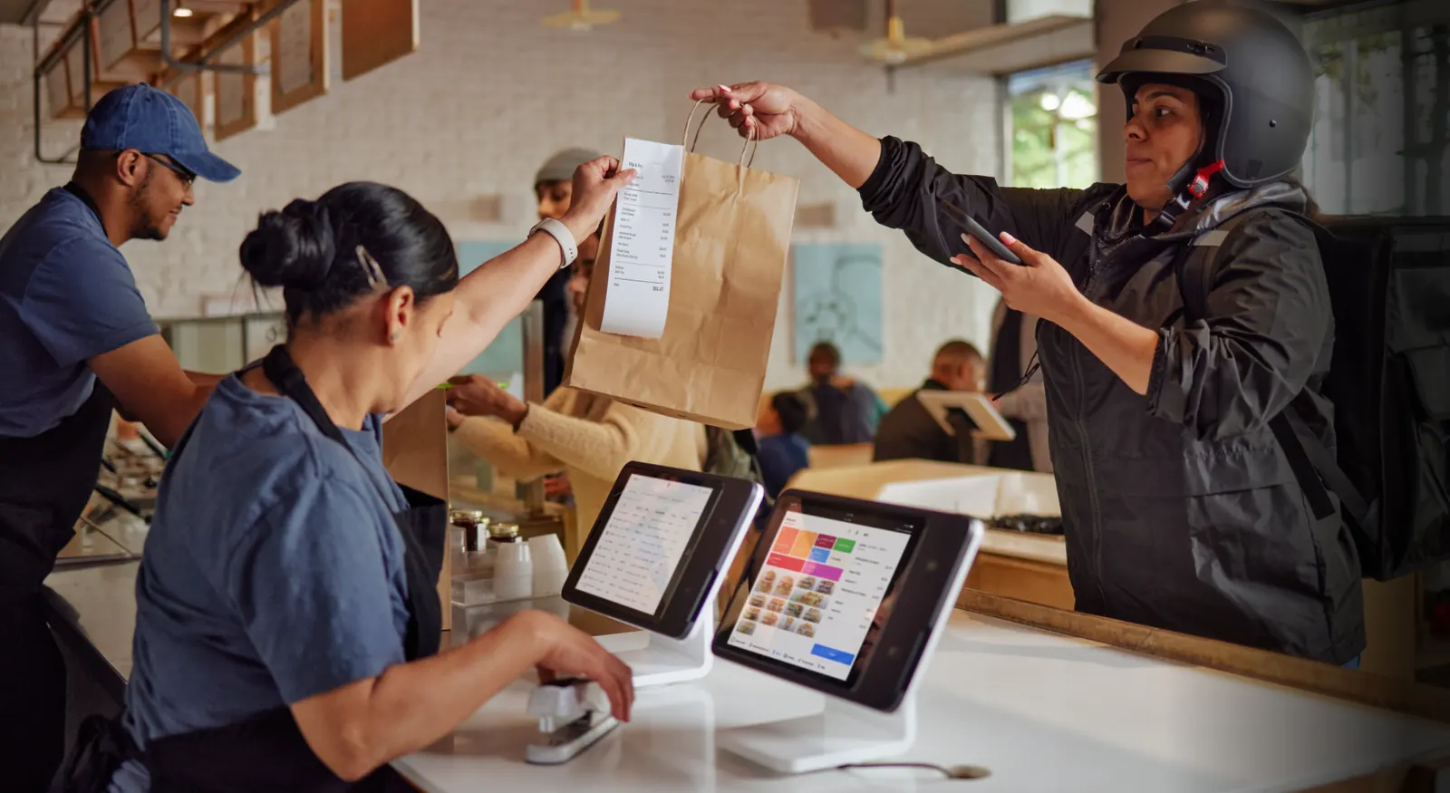 Two restaurant workers work on the register while handing a take-out bag to a delivery person to the right.