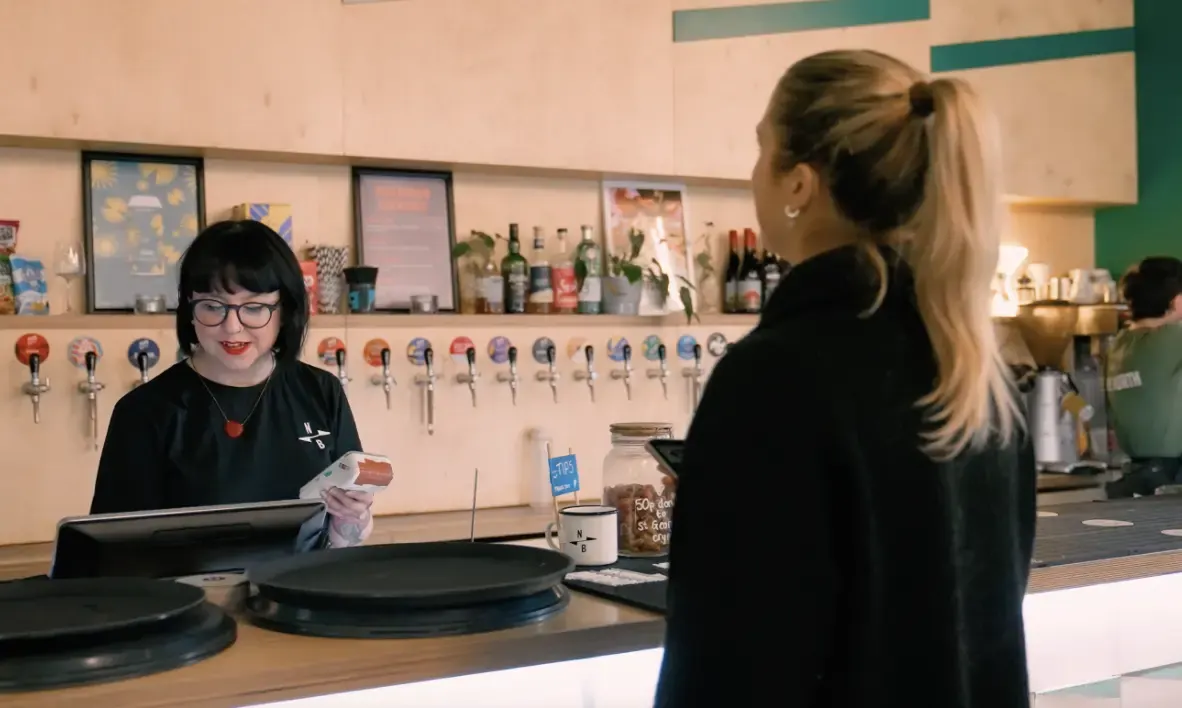 Bar staff member behind a counter with a till, facing a customer